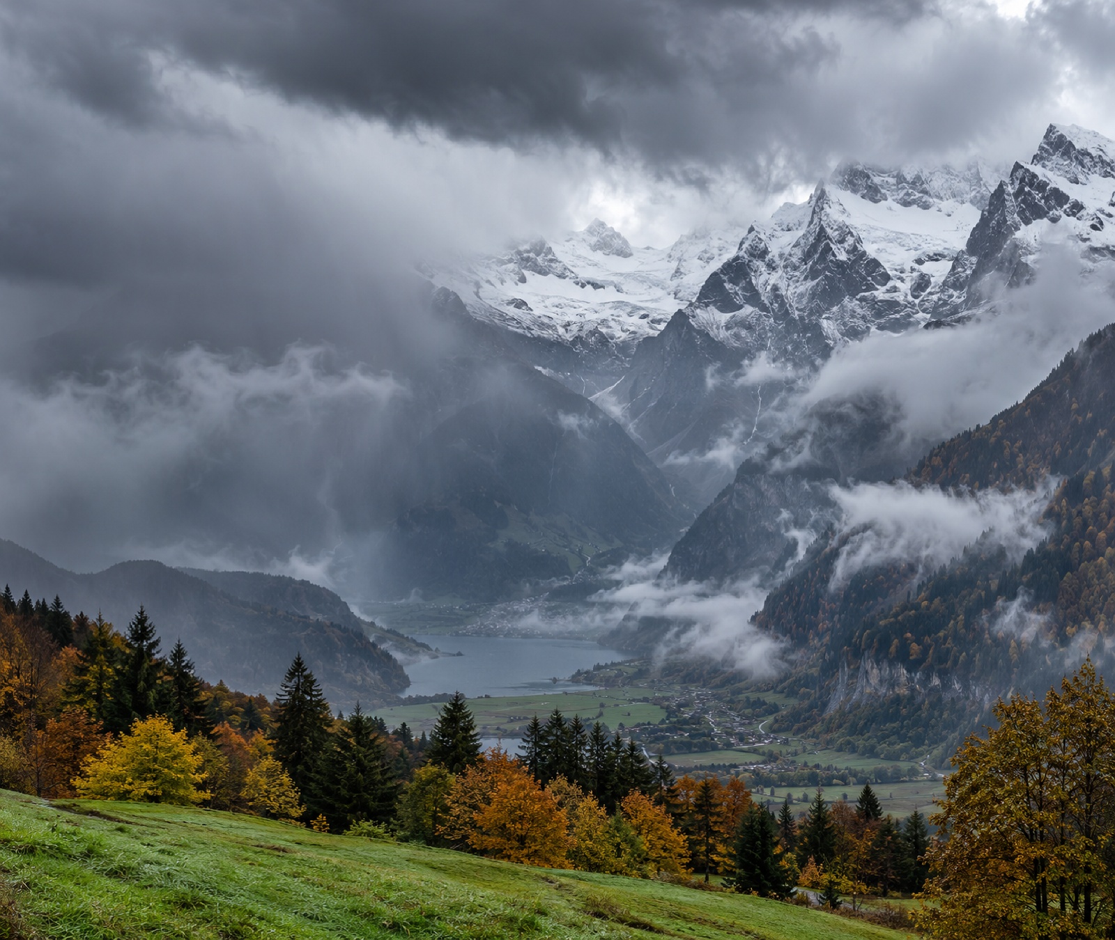 Vallée alpine française sous de gros nuages avec neige sur les hauts sommets et couleurs d’automne plus bas, image d’illustration.