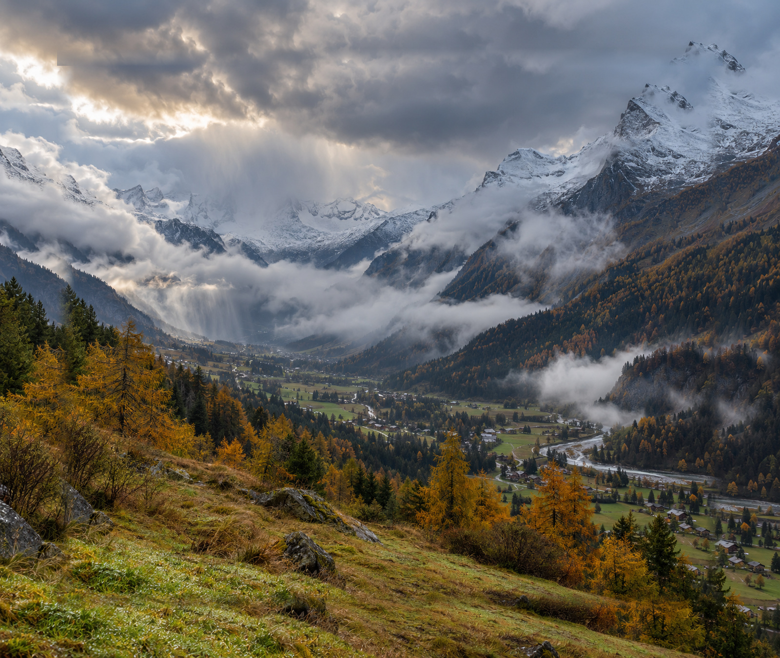 Vallée des Alpes françaises en automne sous des nuages bas et des précipitations, avec neige fraîche sur les hauts sommets.